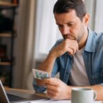 Man in a denim shirt sitting at a desk with a laptop, thoughtfully examining a blister pack of blue pills.