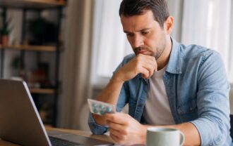 Man in a denim shirt sitting at a desk with a laptop, thoughtfully examining a blister pack of blue pills.